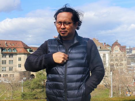 Person wearing a dark vest and jeans standing in front of a hedge and historic buildings under a blue sky.