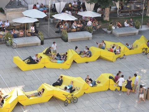 Gelbe Enzi-Sitzmöbel im MuseumsQuartier Wien, Menschen entspannen in der Sonne nahe der Caféterrasse.