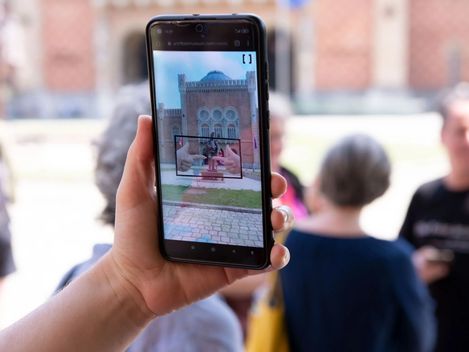 Hand holding smartphone showing a photo of a historic building with people in the background.