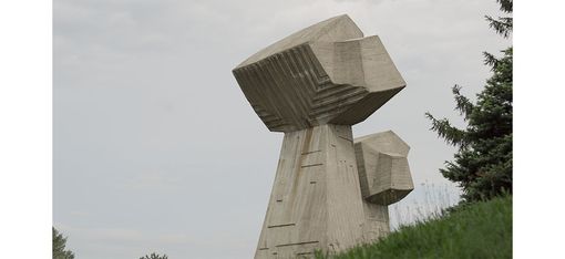 Große, abstrakte Betonplastik mit zwei kubischen Formen auf einem geneigten Hügel vor bewölktem Himmel.