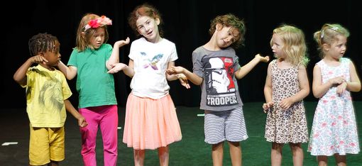 Sechs Kinder in einem scharzen Raum, der in grünes Licht getaucht ist. Die Mädchen und Buben machen unterschiedliche Gesten, zeigen auf das Handgelenk oder in den Raum, drei haben hochgezogene Schultern, angewinkelte Arme und schief gelegte Köpfe, als wollten sie ausdrücken "ich weiß auch nicht, was hier los ist".