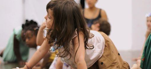 Ein Gruppe von kleinen Kindern und eine erwachsene Person unscharf im Hintergrund, im Vordergrund ein kleines Mädchen, das sich gerade auf allen Vieren vorwärts bewegt, den Kopf nach rechts geneigt.