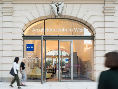 Eine Person in schwarz gekleidet und zwei weitere Personen gehen bei einer großen, verglasten Tür mit Rundbogenfenster vorbei. Darauf steht Architekturzentrum Wien.