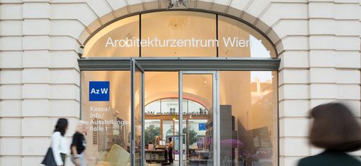 A person dressed in black and two other people walk past a large glass door with a round-arched window. It says Architekturzentrum Wien on it.