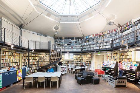 Room with tables and armchairs, with several people, bookshelves with many books in the background. Ceiling with octagonal window.