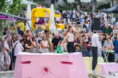 Menschenmenge bei einer Veranstaltung im Freien mit einer Frau, die auf einer pinkfarbenen Betonbarriere tanzt, und einem Mann in weißem Hemd und Mütze neben ihr