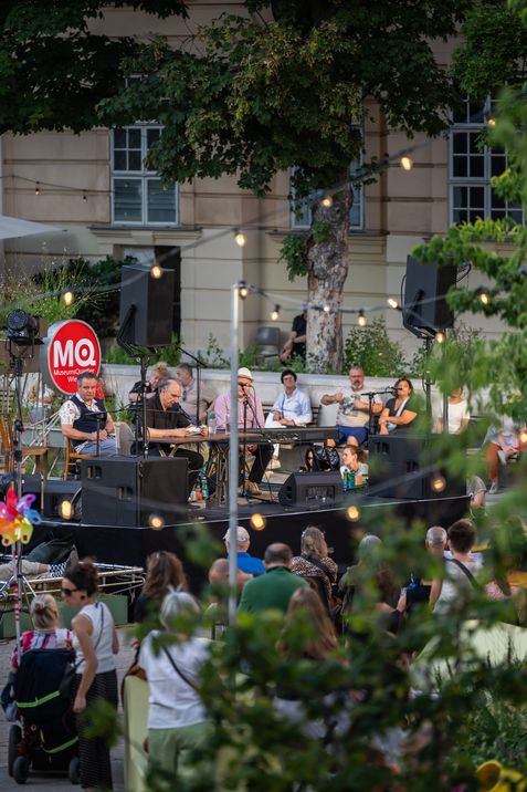 Several musicians seated on an outdoor stage in front of an audience surrounded by trees and string lights with a red MQ logo in the background