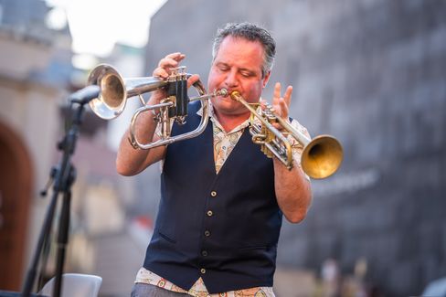 Man wearing vest playing two trumpets simultaneously on an outdoor stage