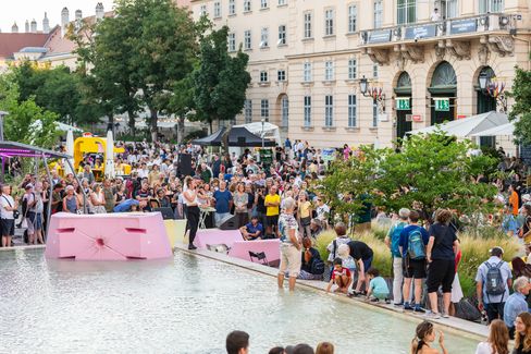 Large crowd gathered around a water basin with pink seating in front of a historic building in daylight