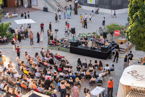 Bird's-eye view of an outdoor event with audience seated in front of a stage and several people walking on a plaza with trees and tents
