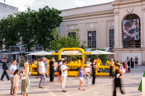People move around a yellow open pavilion with umbrellas on a plaza in front of a historic building with large windows and a banner on the facade