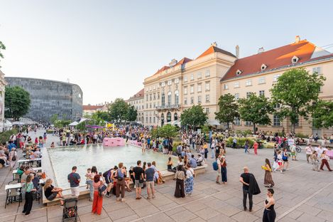 Crowd of people around a large rectangular reflecting pool in front of historic buildings during daylight