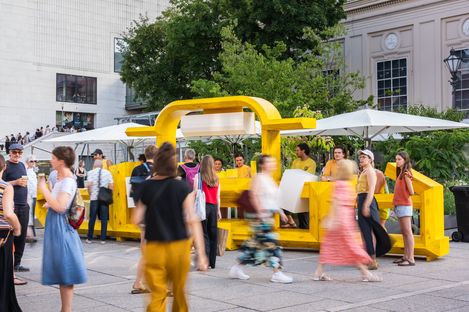 Menschen bewegen sich um eine gelbe, skulpturale Sitzbank mit Sonnenschirmen in einem urbanen Außenbereich mit Bäumen und Gebäuden im Hintergrund