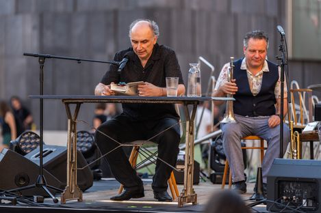 Four people sitting on an outdoor stage with musical instruments and microphones in front of an audience