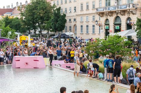 Large crowd gathered around a water basin with pink seating in front of a historic building in daylight