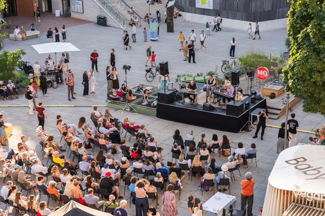 Bird's-eye view of an outdoor event with audience seated in front of a stage and several people walking on a plaza with trees and tents