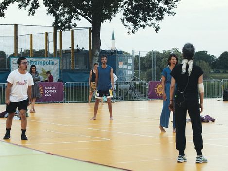 Five people stand on a sports court, one person with a microphone and headphones faces away from the camera while the others look toward them