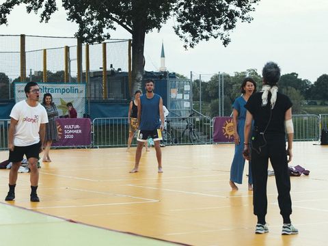 Five people stand on a sports court, one person with a microphone and headphones faces away from the camera while the others look toward them
