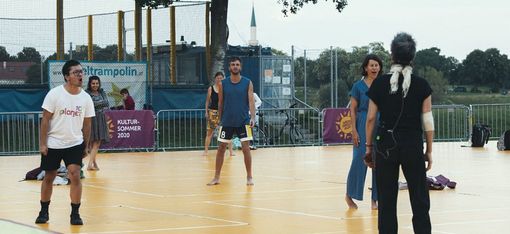 Five people stand on a sports court, one person with a microphone and headphones faces away from the camera while the others look toward them