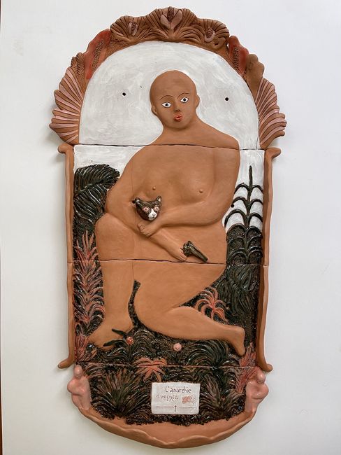 Ceramic sculpture of a naked kneeling person holding a small animal surrounded by plant motifs and a white background.