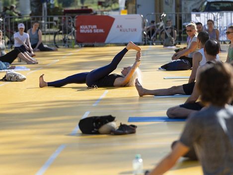 Person lying on back on a mat holding one leg stretched upward surrounded by other people on mats outdoors