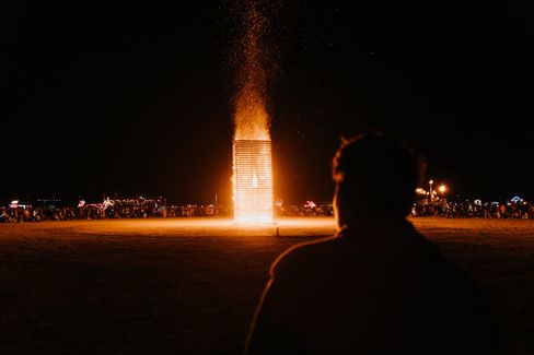 Silhouette of a person in front of a large burning wooden structure on an open ground at night with a crowd in the background.