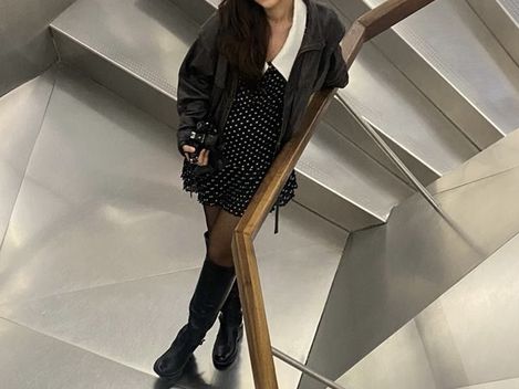 Frau mit dunklem Haar, schwarzem Kleid mit weißen Punkten, schwarzer Jacke und schwarzen Stiefeln steht auf einer modernen Treppe mit Metallstufen und Holzgeländer.