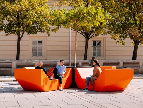 Menschen sitzen und unterhalten sich auf orangefarbenen Enzis im MuseumsQuartier, umgeben von Bäumen und historischer Architektur