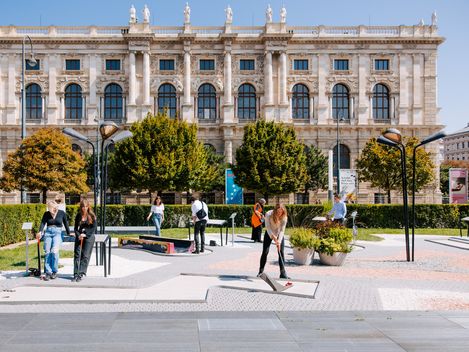 Mehrere Personen spielen Minigolf auf einer modernen Anlage vor einem historischen Gebäude mit symmetrischer Fassade.