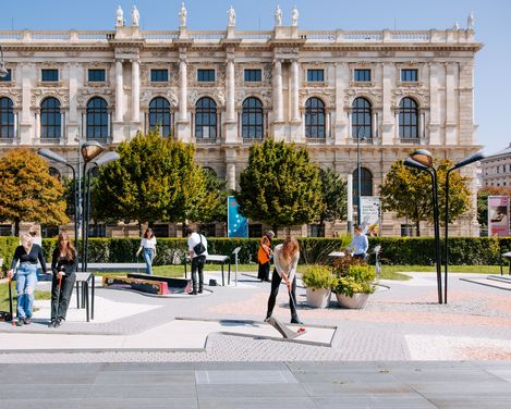 Mehrere Personen spielen Minigolf auf einer modernen Anlage vor einem historischen Gebäude mit symmetrischer Fassade.