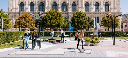 Multiple people play mini golf on a modern course in front of a historic building with a symmetrical facade.