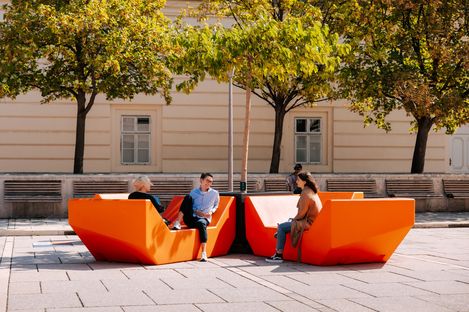 People sitting and talking on orange Enzis at MuseumsQuartier, surrounded by trees and historic architecture