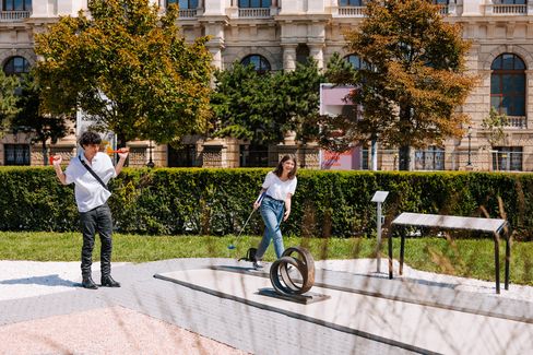 Two people stand and walk on an outdoor mini-golf course with metal obstacles in front of a historic building.