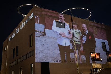 Projektion eines Fotos mit drei Personen auf die Fassade eines Gebäudes mit der Aufschrift "Artists for Ukraine".