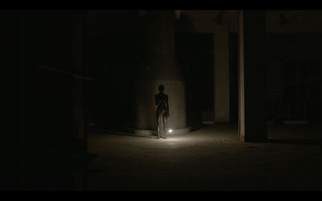 A single person stands in a dark, spacious room in front of a column, illuminated by a light on the floor.