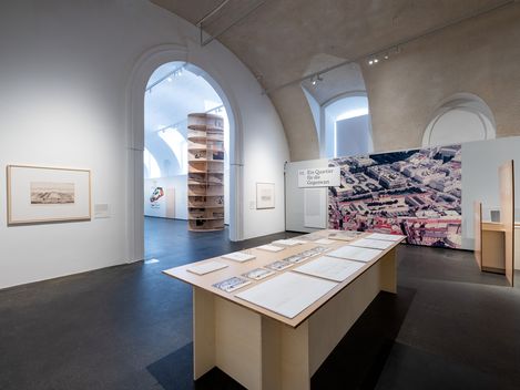 Exhibition room with white walls, large arched windows, a long table displaying drawings and photographs, and a large aerial photograph on the wall