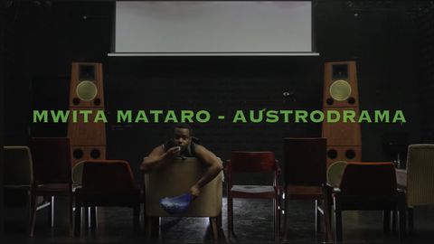 Mann sitzt auf einem Sessel in einem dunklen Raum mit mehreren Stühlen und zwei großen Lautsprechern, Text "MWITA MATARO - AUSTRODRAMA" in grüner Schrift über ihm.