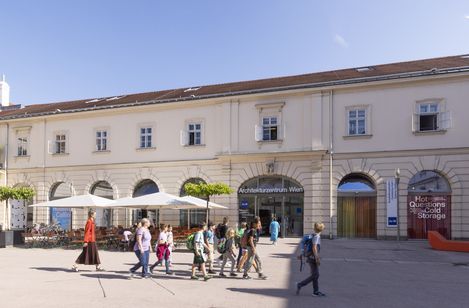Several people are walking in one direction across a courtyard with a paved floor. Behind them is a building with large arched windows and large parasols with tables and chairs
