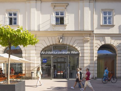 Ein paar Menschen gehen vor einer großen Tür mit Rundbogenfenstern vorbei, auf der "Architekturzentrum Wien" drauf steht. Eine Frau in einem türkisen Kleid schiebt ein Fahrrad.