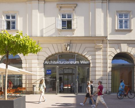 A few people walk past a large door with arched windows that says “Architekturzentrum Wien”. A woman in a turquoise dress pushes a bicycle.