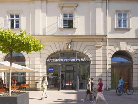 Ein paar Menschen gehen vor einer großen Tür mit Rundbogenfenstern vorbei, auf der "Architekturzentrum Wien" drauf steht. Eine Frau in einem türkisen Kleid schiebt ein Fahrrad.