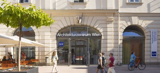 Ein paar Menschen gehen vor einer großen Tür mit Rundbogenfenstern vorbei, auf der "Architekturzentrum Wien" drauf steht. Eine Frau in einem türkisen Kleid schiebt ein Fahrrad.
