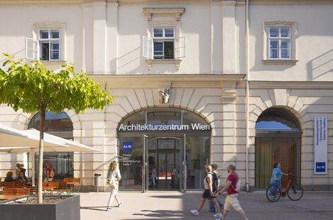 A few people walk past a large door with arched windows that says “Architekturzentrum Wien”. A woman in a turquoise dress pushes a bicycle.
