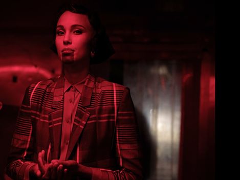 Woman wearing a plaid blazer and shirt standing against a red background holding her hands in front of her.