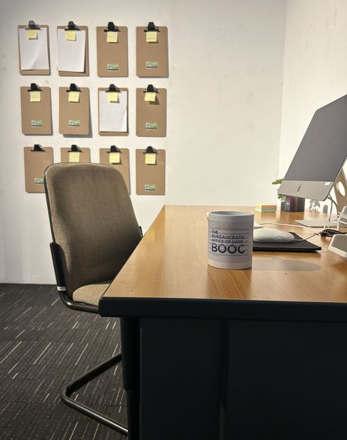 Schreibtisch mit Computer, Tasse („Bureaucratic Office of Care“) und Stuhl; an der Wand hängen Klemmbretter mit Notizen.