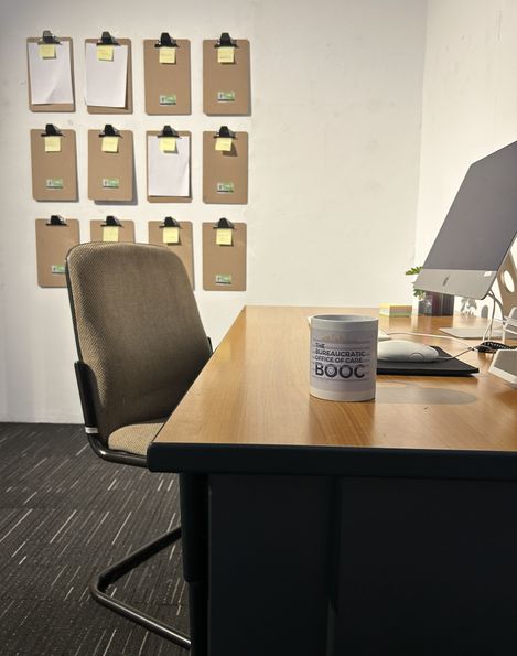 Schreibtisch mit Computer, Tasse („Bureaucratic Office of Care“) und Stuhl; an der Wand hängen Klemmbretter mit Notizen.