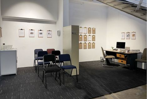 Installationsansicht eines raumartigen Set-ups mit Stuhlreihen, Schreibtisch und Wänden voller Klemmbretter und Dokumente.