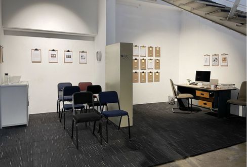 Installationsansicht eines raumartigen Set-ups mit Stuhlreihen, Schreibtisch und Wänden voller Klemmbretter und Dokumente.