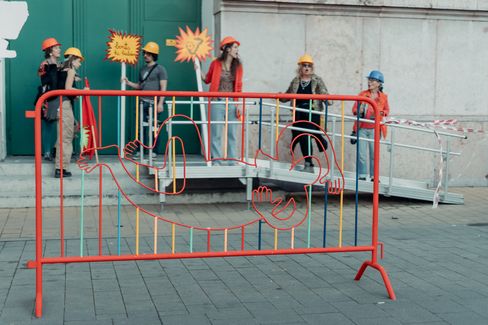 Six people wearing construction helmets stand behind an orange construction barrier with a colorful wire sculpture shaped like a reclining person.