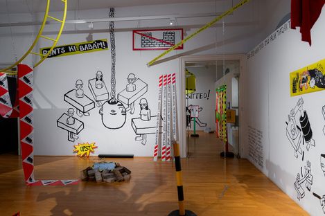 Art installation with black-and-white wall drawings of construction workers at desks, a large wrecking ball hanging from a chain, suspended chains, and colorful ribbons in a room with wooden floor.